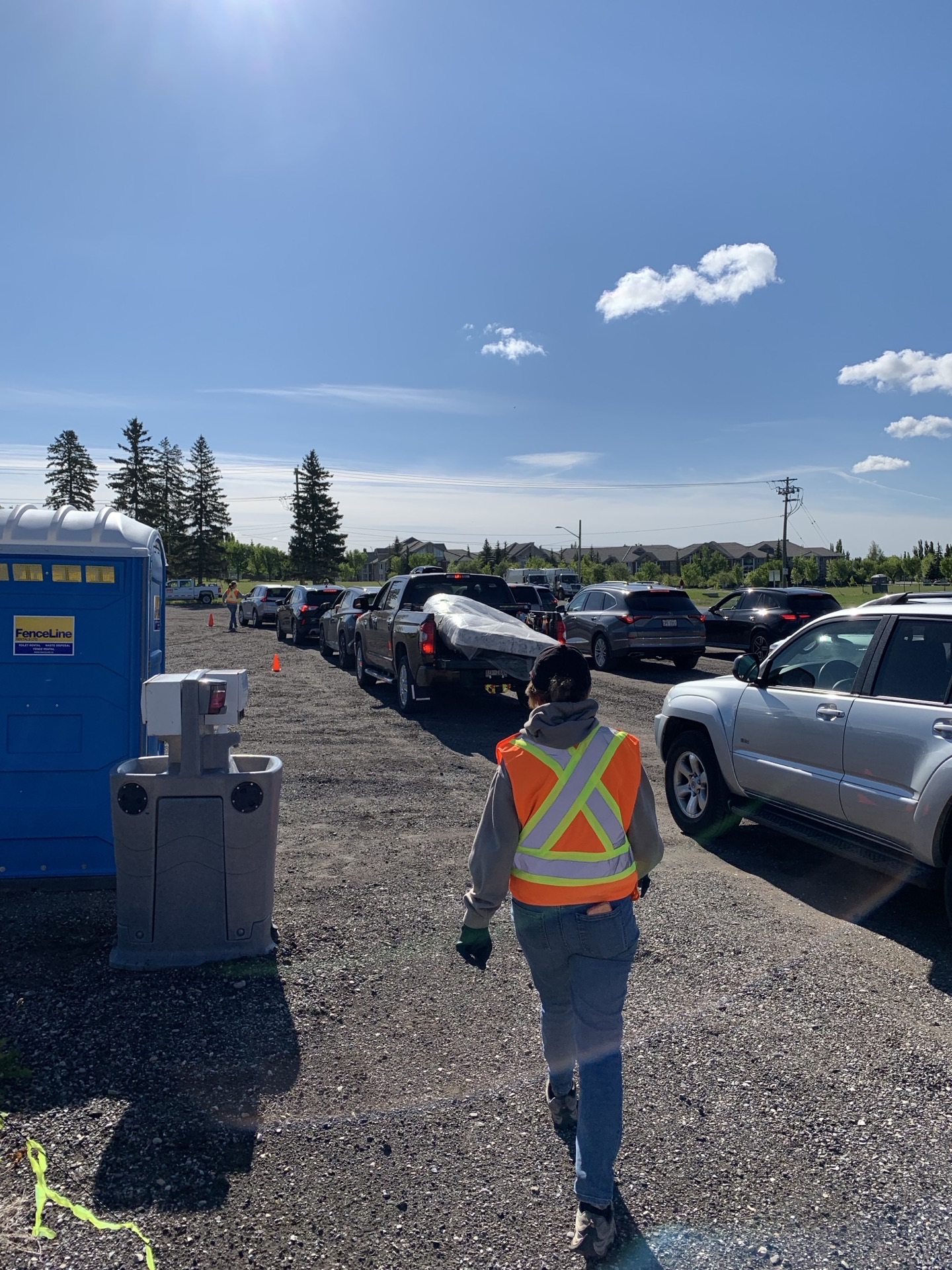 A volunteer in a high-vis vest walking down a line of cars queued for drop-off, porta-potty and traffic cones along the lane.