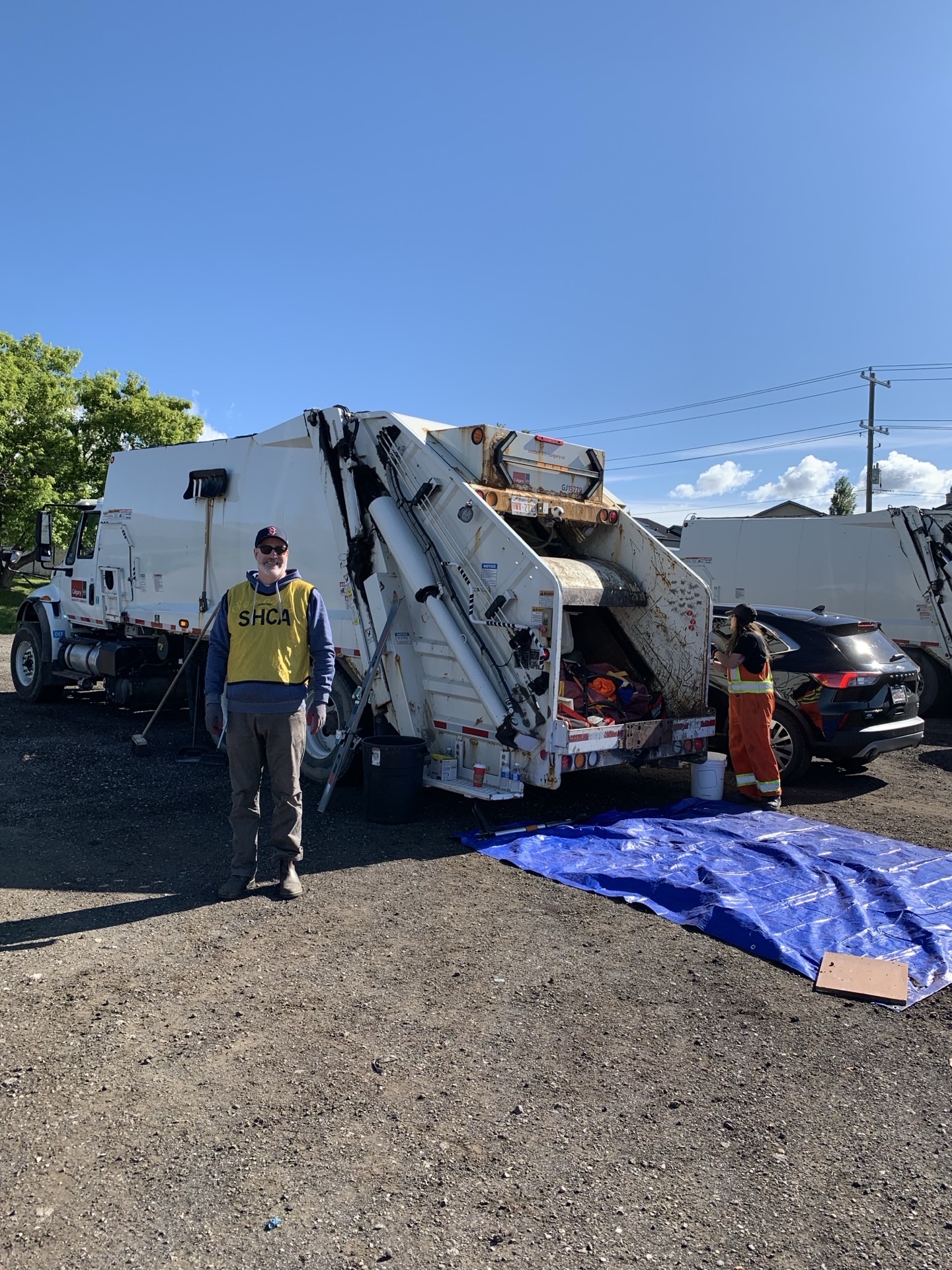 A solo SHCA-vested volunteer standing in front of the garbage trucks, blue tarp staged behind.