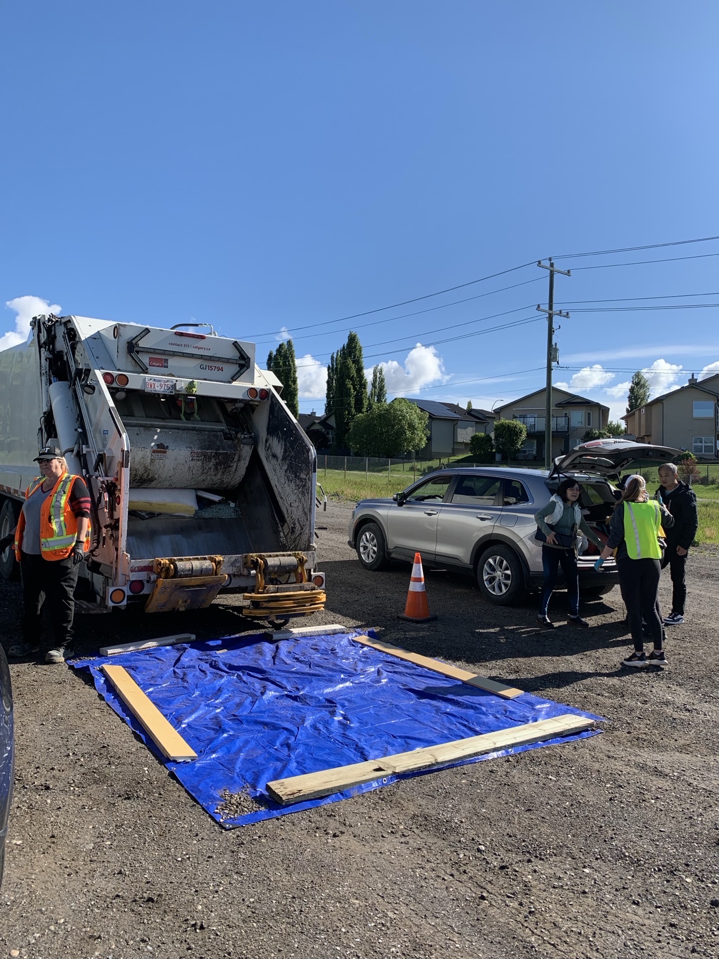 Back of the garbage truck mid-load, a resident sorting items out of their hatchback, blue tarp staged for the drop.