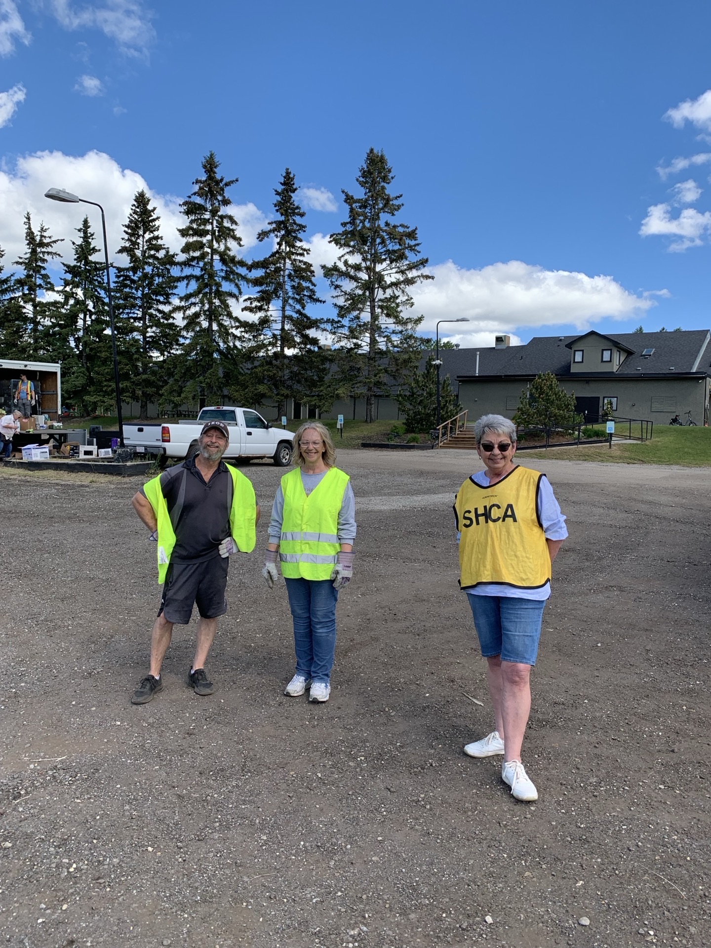 Three volunteers in yellow SHCA safety vests standing on the staging gravel, evergreens and houses behind them.