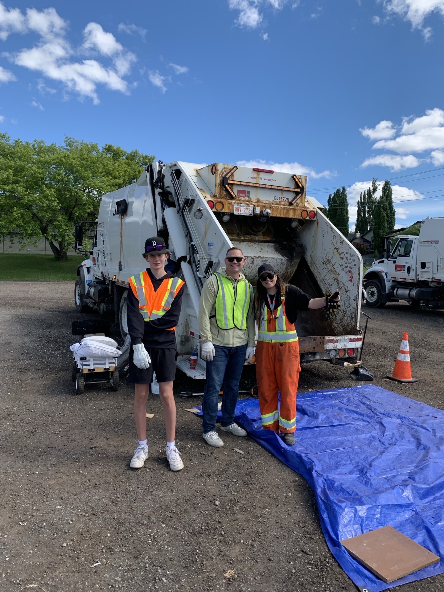 A younger crew of three volunteers in front of the garbage truck, orange traffic cones along the tarp.
