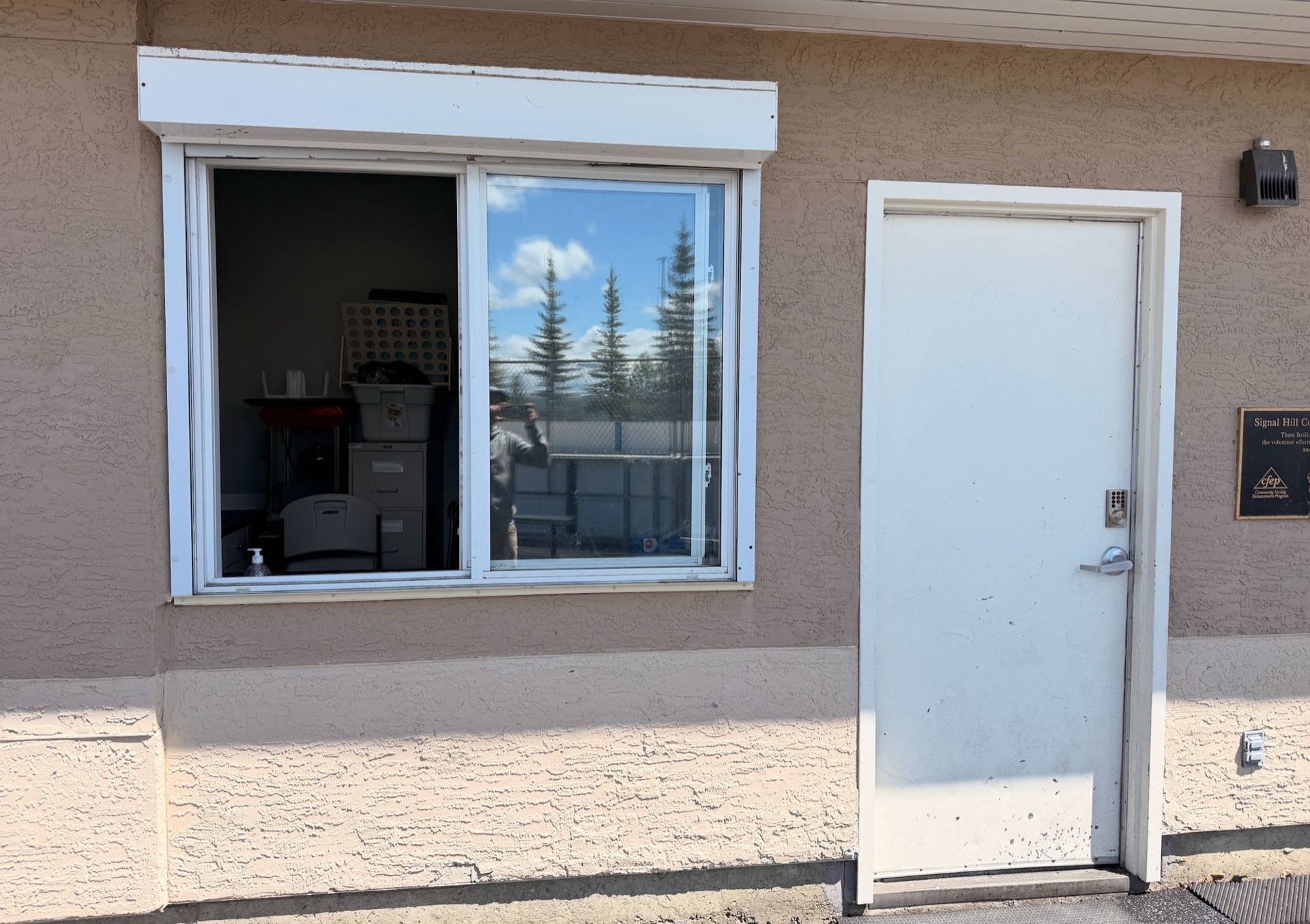 The SHCA kids' vendor window — exterior view showing the open sliding service window with white roll-up shutter housing above, alongside the keypad-locked side door for setup access.