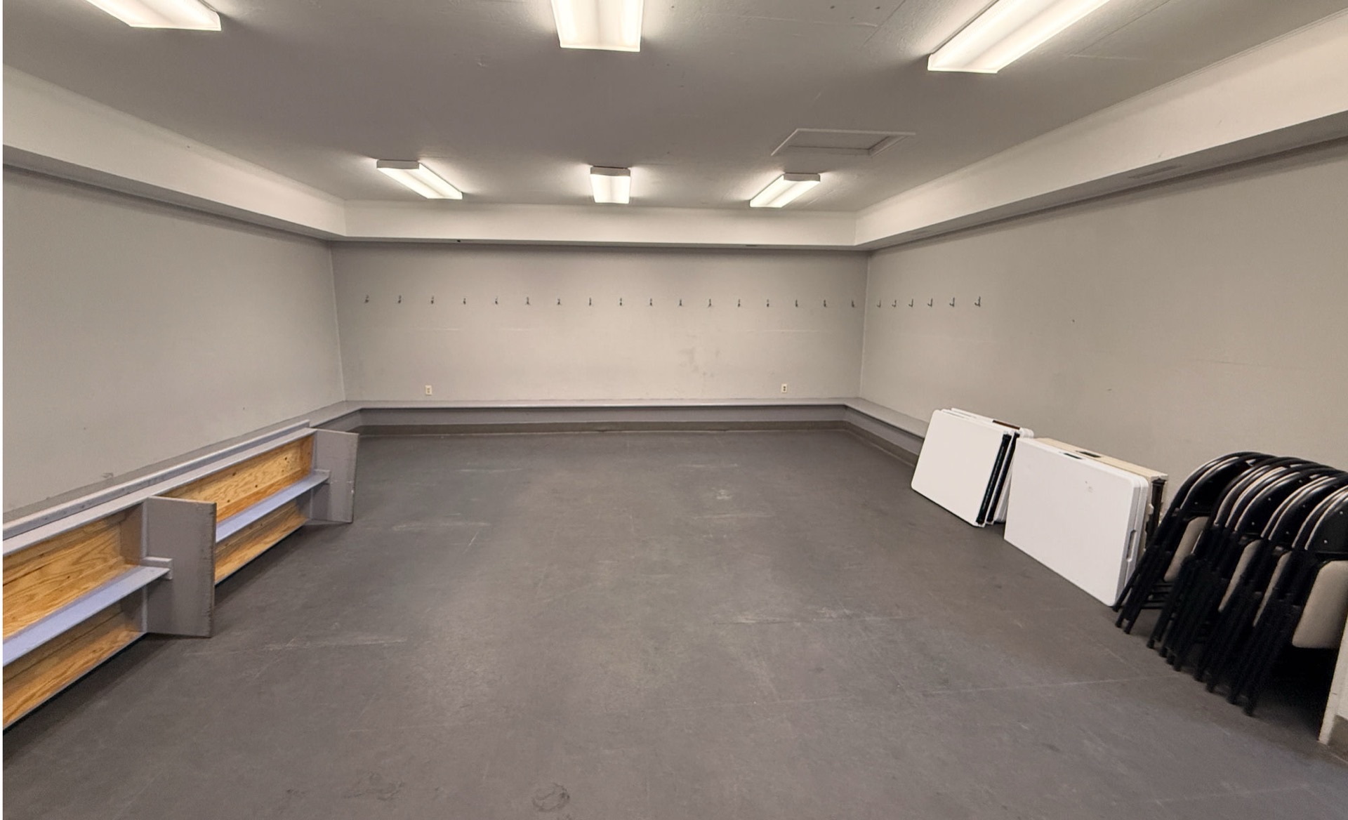 The SHCA indoor multi-purpose room — clean grey-floor space under overhead fluorescent lighting, wooden bench along the left wall, folding tables and stacked chairs ready against the right wall, long row of coat hooks across the back.