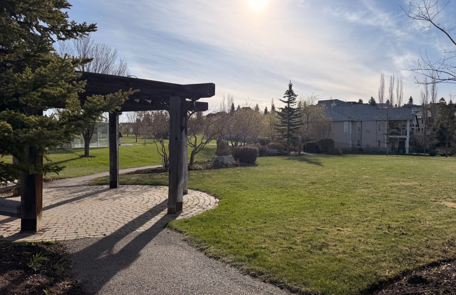 The Signal Hill park gazebo — a dark-wood pergola on a circular brick patio at the edge of an open lawn, gravel pathway curving through, evergreens and Signal Hill homes in the late-afternoon golden sun.