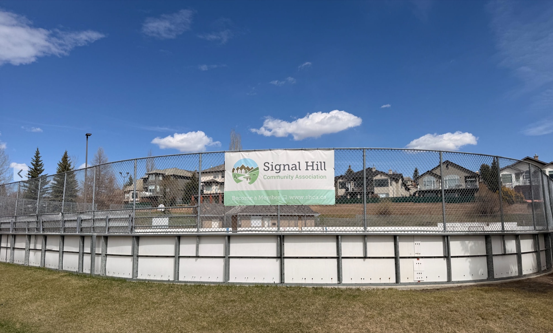 The Signal Hill rink from outside the chain-link fence — white perimeter boards around the slab, SHCA 'Become a Member' banner mounted on the fence, Signal Hill homes behind, blue sky with scattered cloud.