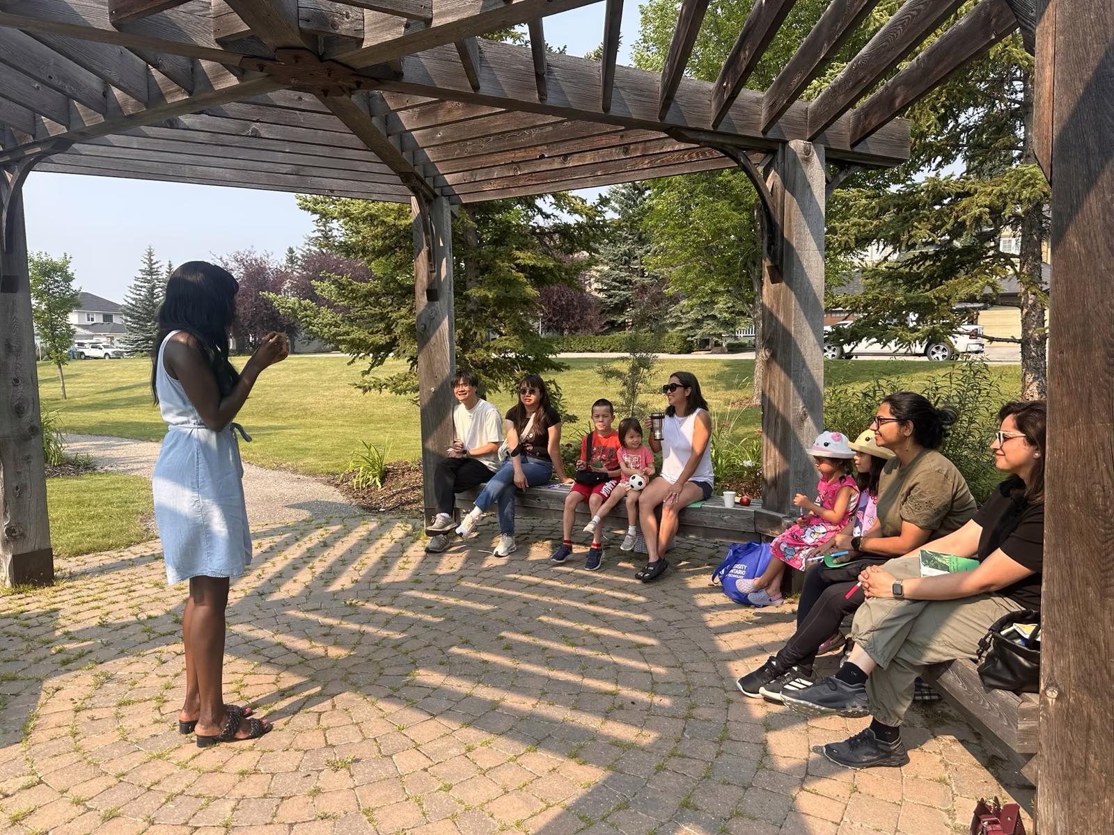 Iman standing in front of the group under the gazebo, leading the conversation while neighbours listen from the benches