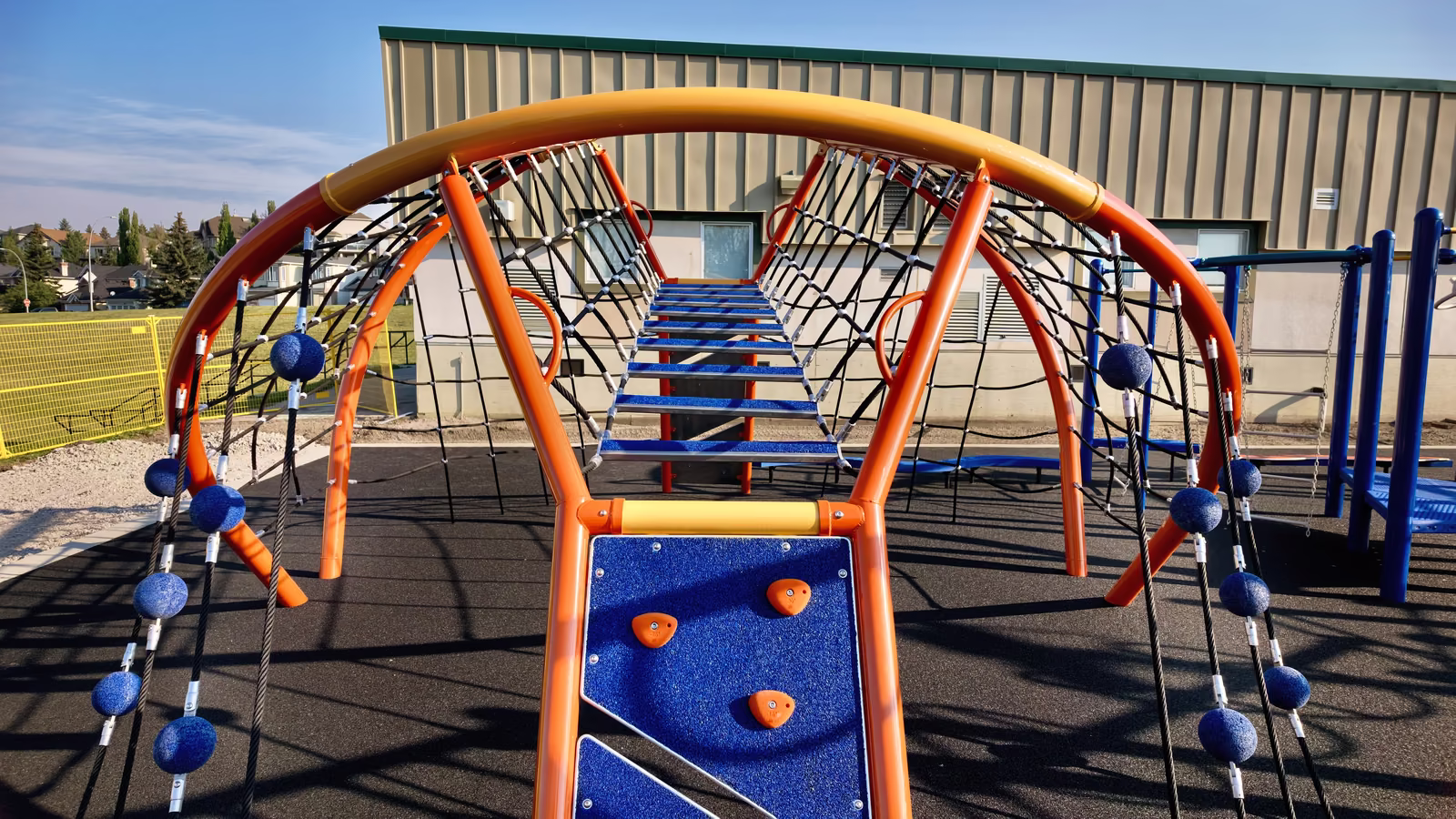 A large orange arched rope climbing structure with a net bridge through the centre and a blue rock wall at the entrance