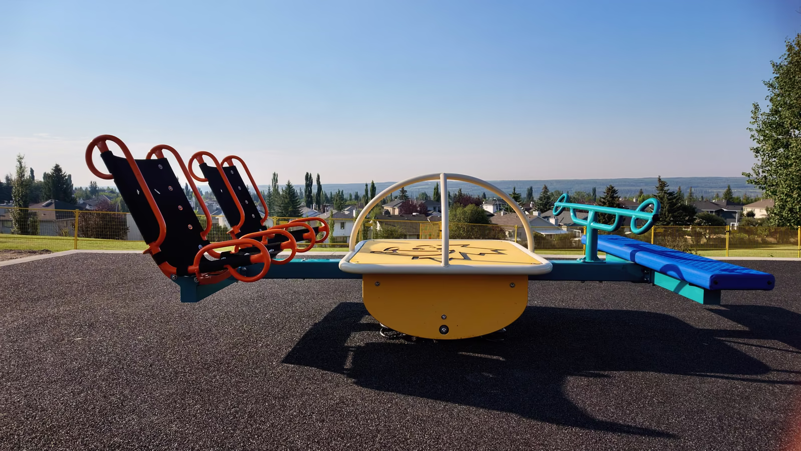 A red multi-seat rocker, a yellow spinner and a teal teeter totter at the edge of the playground, with rooftops and the city skyline in the distance