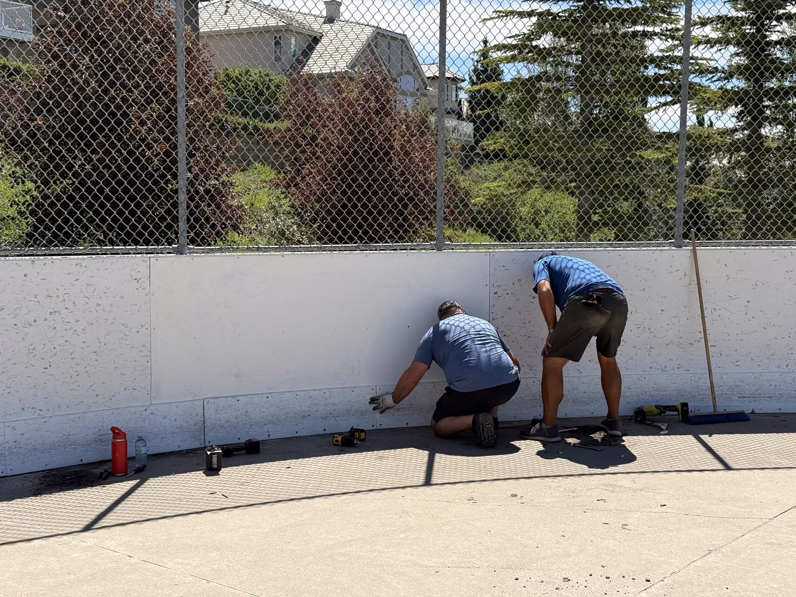 Two volunteers in blue kneeling at the base of the white rink boards, tools laid out