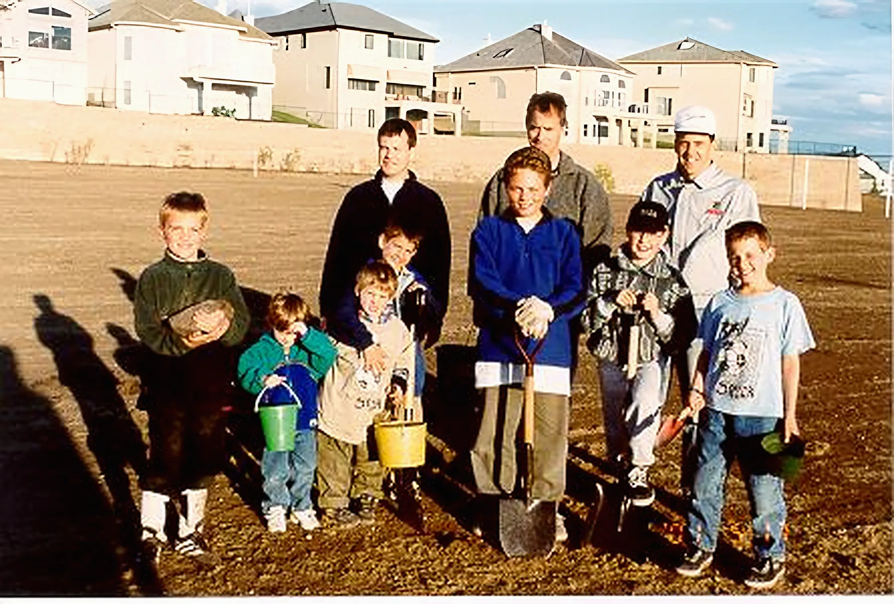 Three dads and eight kids stand on a bare, freshly turned field holding buckets, shovels and rocks, with a row of new two-storey houses along the ridge behind them. May 2001.